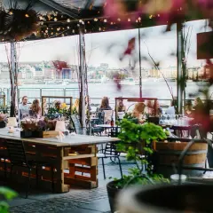 En mysig fotografisk veranda med utsikt över vattnet och stadens silhuett. Människor sitter vid borden och njuter av atmosfären.