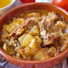 Guiso de cap i pota en cazuela de barro, con patatas y trozos de carne, acompañado de ingredientes frescos.