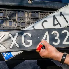 Wymiana tablic rejestracyjnych. Jedna tablica z numerem KXG 4325 jest przykręcana, druga z numerem C414 jest trzymana w ręku.