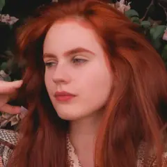 Chica con vibrante pelo pelirrojo, rodeada de flores y follaje, con las manos jugando con su cabello.