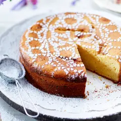 Ein saftiger Joghurt Kuchen, mit Puderzucker bestäubt und einem Stück herausgeschnitten. Daneben liegt ein kleines Sieb.