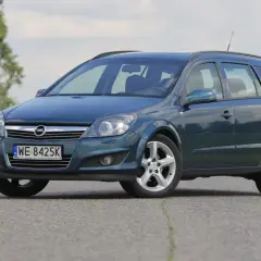Granatowy Opel Astra Cosmo w wersji kombi stoi na asfaltowej drodze, otoczony zielenią i drzewami.