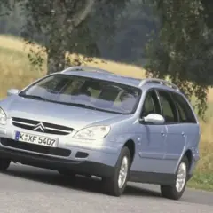Jasnoniebieskie kombi Citroen na zakręcie drogi, z drzewami i polem w tle.