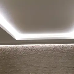 Illuminazione indiretta su velette in cartongesso, che creano un effetto soffuso su una parete in pietra chiara.