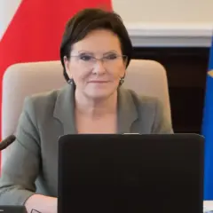Ewa Kopacz, premier, siedzi przy stole z laptopem, obok flagi Polski i Unii Europejskiej.