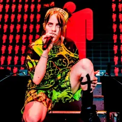 Billie Eilish na koncercie, w neonowym stroju, z mikrofonem w dłoni. Tło zdobią czerwone sylwetki ludzi.