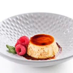 Delicioso flan de queso fresco con caramelo líquido, decorado con frambuesas y menta en un plato blanco texturizado.