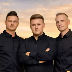Trzech mężczyzn w czarnych koszulach, tworzących zespół Diadem, pozuje na tle zachodzącego słońca.