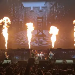 Płomienie buchają na scenie, gdzie grają zespoły rockowe. W tle widać zamek i gilotynę.