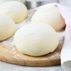 Kulki ciasta, idealne ciasto na pizzę, posypane mąką, leżą na desce. Obok różowy ręcznik.