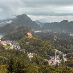 Schlechtes Wetter trübt den Urlaub. Ein gelbes Schloss thront auf einem bewaldeten Hügel über einem Dorf am See.