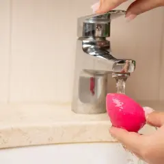 Mano limpiando esponja de maquillaje rosa bajo un grifo. Aprende como limpiar las esponjas de maquillaje para un mejor acabado.