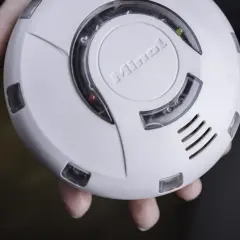 Ein weißer enocean rauchmelder mit dem Schriftzug "Minol" wird von Fingern gehalten. Ein rotes und ein gelbes Licht leuchten.
