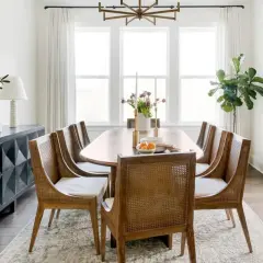 A spacious dining room with a large oval table, cane-backed chairs, and a modern sideboard. The dining room width is emphasized by the large windows and glass doors.