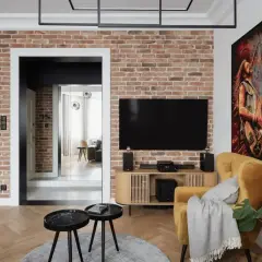 Nowoczesny salon z ceglaną ścianą, na której subtelne oświetlenie kamienia podkreśla jego fakturę. Obok telewizor i żółty fotel.