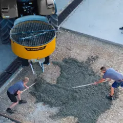 Zwei Arbeiter verteilen frischen Beton, der aus einem gelben Zwangsmischer einer Mammut-Maschine fließt. Ein dritter Arbeiter glättet den Beton.