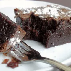 Un morceau de gâteau chocolat, saupoudré de sucre glace, est sur une assiette blanche. Une fourchette est prête à déguster ce délice.
