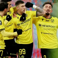 Piłkarze w żółtych koszulkach świętują bramkę. Fani spekulują o plotkach transferowych Widzewa, a drużyna cieszy się sukcesem.