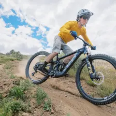 A mountain biker shreds a dusty trail on a Yeti MTE bike, kicking up dirt under a cloudy sky.