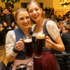 Zwei fröhliche Frauen in Tracht halten je ein großes, gestauchtes Bier. Im Hintergrund ist eine belebte Gaststube zu sehen.