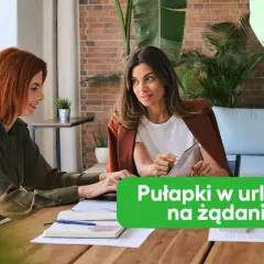Dwie kobiety rozmawiają przy biurku, jedna pokazuje coś na tablecie.