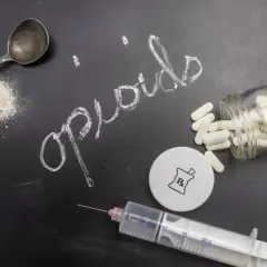 Na czarnej tablicy napis "opioids" kredą. Obok łyżka z proszkiem, strzykawka, fiolka z kapsułkami i symbol apteczny.