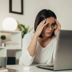 Zestresowana kobieta z okularami masuje skronie, patrząc na ekran laptopa. Brak odpowiedzi na reklamację pogłębia jej frustrację.