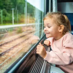 Uśmiechnięta dziewczynka patrzy przez okno pociągu. Zastanawia się, od ilu lat można jeździć samemu pociągiem, marząc o podróżach.