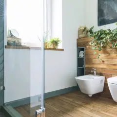 Nowoczesna łazienka z bidetem, który służy do higieny intymnej. Obok stoi miska WC. Drewniane panele i rośliny tworzą przytulny klimat.