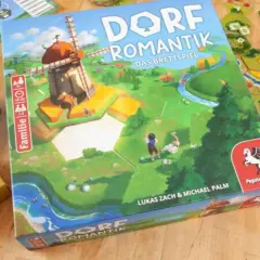 Das Familienspiel des Jahres "Dorfromantik" mit seinen bunten Spielsteinen und der Hauptbox, die eine idyllische Landschaft mit Windmühle zeigt.