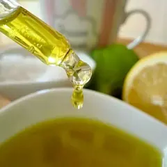 Tropfen eines goldenen Öls fallen aus einer Pipette in eine Schale. Zitronen und Limetten im Hintergrund deuten auf natürliche Inhaltsstoffe für Kosmetik hin, wie z.B. Zitronensäure.