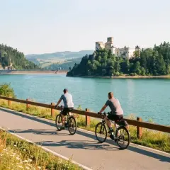 Dwóch rowerzystów na ścieżce Velo Dunajec Czorsztyn, zamek w tle i turkusowa tafla jeziora.