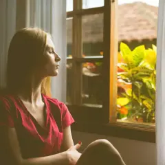 Kobieta w czerwonej bluzce siedzi przy oknie, zamyślona. Szuka sposobów, jak radzić sobie z lękiem, szukając spokoju w naturze za oknem.