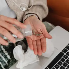 Dłoń z kapsułkami leków, obok laptop i chusteczki.