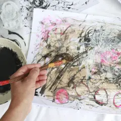 A hand holds a brush, adding bold strokes to an abstract drawing. Black ink splatters a palette beside the energetic artwork.