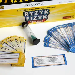 Pudełko gry "Ryzyk Fizyk" i rozłożone karty pytań i odpowiedzi. Czasomierz piaskowy czeka na rozpoczęcie rundy.