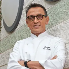 Bruno Barbieri, chef stellato Michelin, sorride con le braccia conserte davanti a un muro di pietra.