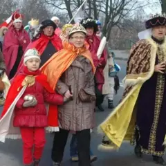 Kolorowe stroje Trzech Króli na ulicznym orszaku. Dziecko w czerwonym płaszczu i koronie, obok dorosłe osoby w bogatych szatach.