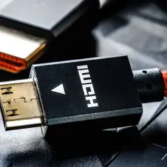 Złote wtyczki HDMI, jedna z nich z czarną obudową i napisem HDMI, połączone z czerwonym, plecionym kablem.