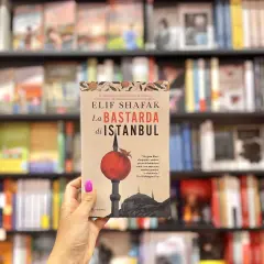 Mano tiene "La Bastarda di Istanbul", un libro che esplora le storie di Istanbul, tra scaffali pieni di libri.