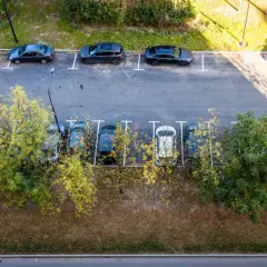 Widok z góry na parkowanie samochodów dostawczych na osiedlu. Kilka aut stoi na wyznaczonych miejscach, otoczonych zielenią i drzewami.