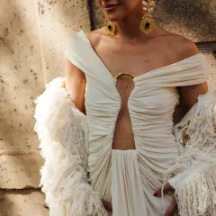 Elegancka sesja fashion: modelka w kremowej sukni z wycięciem i futrzanym bolerkiem, ozdobiona złotą biżuterią.