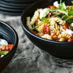 Zwei Schüsseln mit Nudelsalat mit getrockneten Tomaten, Pinienkernen und Rucola. Ein Löffel liegt in der linken Schüssel.