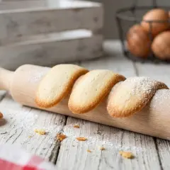 Biscotti lingue di gatto, dorati e spolverati di zucchero a velo, adagiati su un mattarello in legno.
