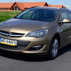 Brązowy Opel Astra J automat w wersji kombi jedzie po drodze. W tle widać domy z czerwonymi dachami.