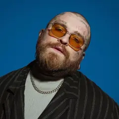 Teddy Swims w okularach przeciwsłonecznych i z tatuażami na głowie, na niebieskim tle.
