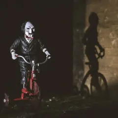 Lalka Billy z Piły na czerwonym trójkołowcu, przypominająca scenę z najlepszych horrorów. Cień na ścianie dodaje grozy.