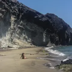 Una mujer descansa en la arena de la cala carbon, con imponentes acantilados de roca volcánica a su espalda.