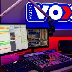 Studio radiowe VOX FM Warszawa. Komputery z listą utworów, mikser, mikrofon i logo stacji.
