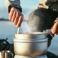 Person lagar mat utomhus, en del av mat vandring. Ångande kastruller och en mugg i metall.